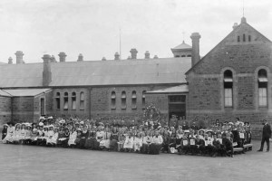 View taken in the yard at the back of Kapunda Primary School, with the children posing for the photographer. A number of teachers are also included. It appears to be a special occasion - there is a large wreath, a number of boys in uniform (two holding rifles), several of the children are holding posters, and all appear to be wearing their Sunday best. DATEca.1900